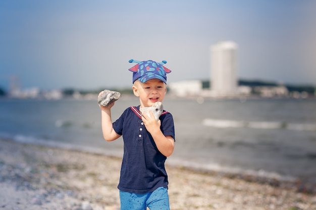 Happy three-year-old child stands on pebbles on the sea beach and holding stones in hands