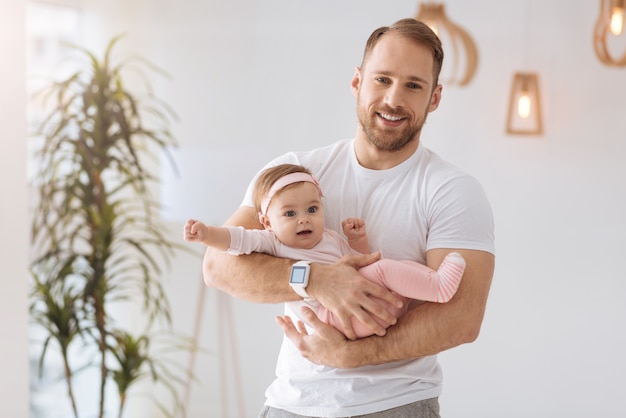 一緒に幸せな時間。家に立って、前向きな感情を表現しながら赤ちゃんを腕に抱いて運動する魅力的な幸せな父