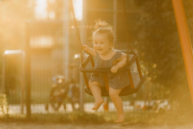 A happy toddler baby girl in a dress on a swing on the warm summer evening