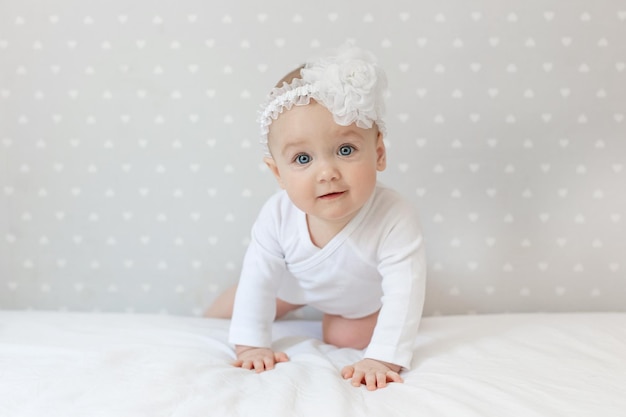 Happy toddler baby girl in a white bodysuit crawls on the bed at home and looking at the camera with smile