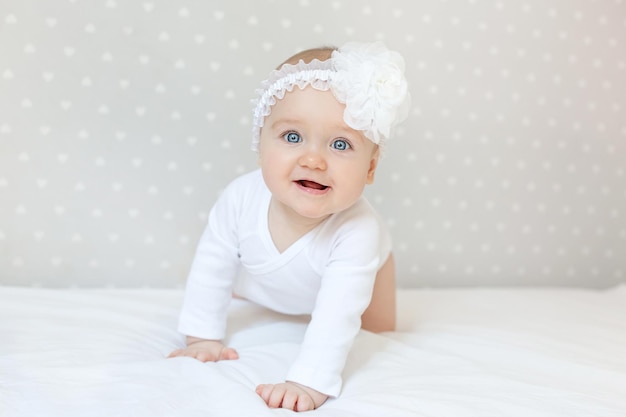 Photo happy toddler baby girl in a white bodysuit sitting on bed at home and looking at the camera with smile. copy space for text