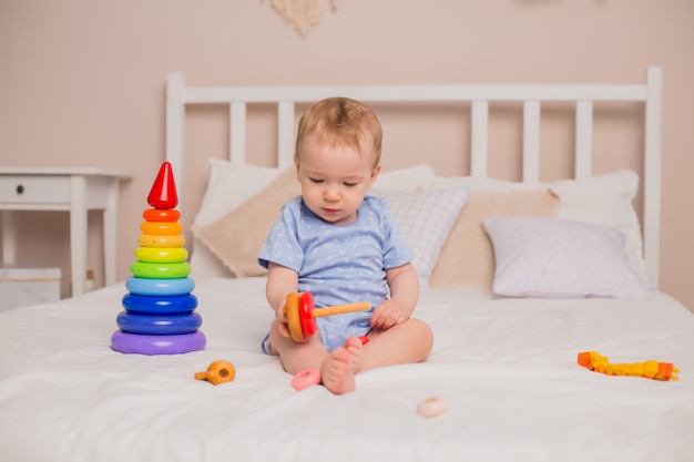 Il bambino felice in tuta blu si siede a casa sul letto che gioca con i giocattoli di sviluppo