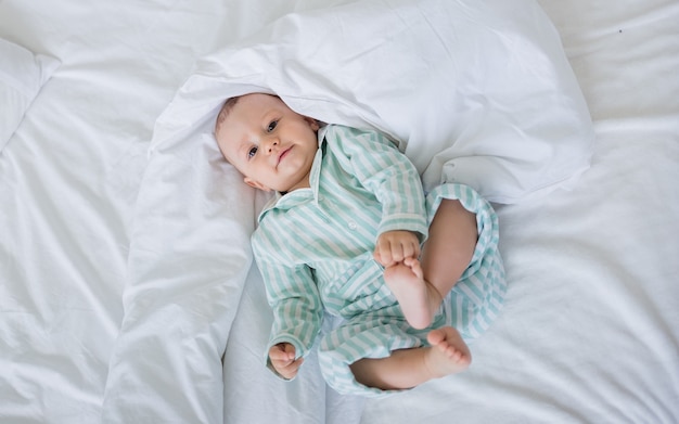 A happy toddler boy in striped pajamas lies on a bed with white linens on a white surface with space for text