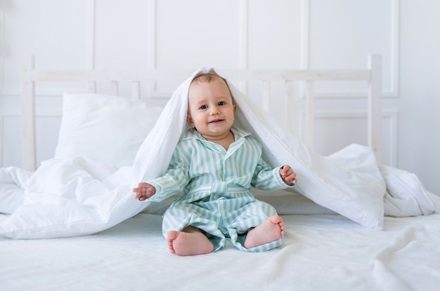 A happy toddler boy in striped pajamas sits on a bed with white linens on a white surface with space for text