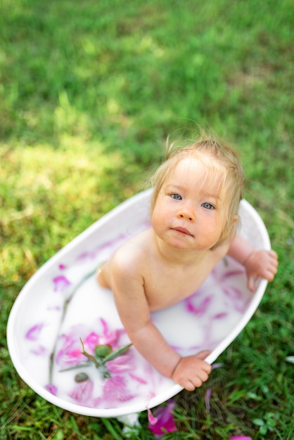 Happy toddler girl takes a milk bath with petals. Little girl in a milk bath . Bouquets of pink peonies. Baby bathing. Hygiene and care for young children.