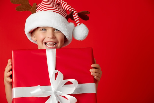 A happy toddler in a striped hat with deer cones holds a large red gift box