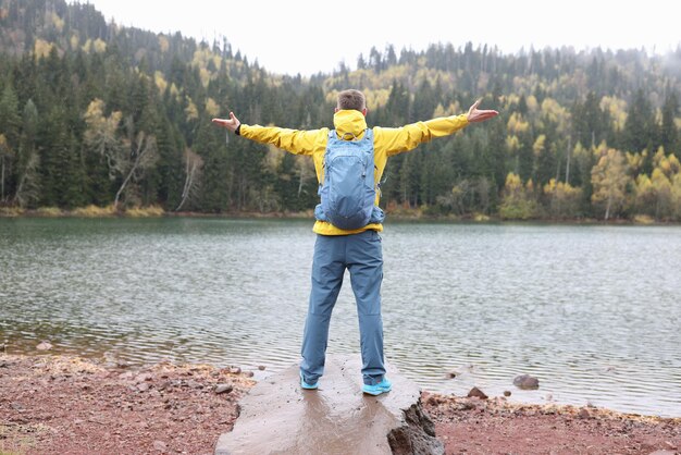 Happy tourist stands with open arms on rock by lake and looks at hills travel outdoor adventure