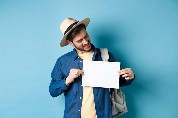 Happy tourist in straw hat smiling and looking at piece of paper for your logo, standing on blue background.