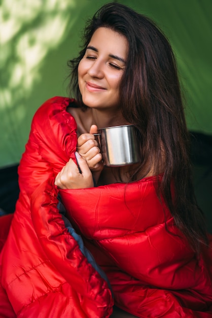 幸せな観光客の女性がテントに座ってお茶を飲んでいます。テントでのキャンプ-キャンプ中の少女。寝袋に入れて、熱いお茶を飲む女性観光客。キャンプ場での朝。ポートレート