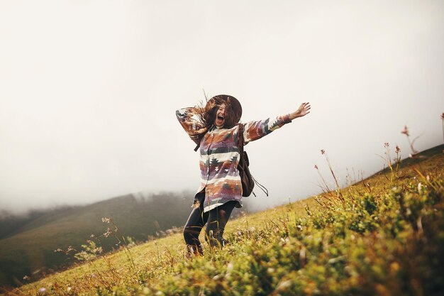 Foto felice viaggiatore hipster ragazza in cappello che salta con lo zaino e sorride nelle montagne nello spazio delle nuvole per il testo incredibile momento emotivo atmosferico viaggio e concetto di voglia di viaggiare