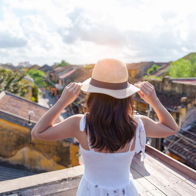 ベトナムのホイアンの古代の町を旅する幸せな旅行者で、ルーフトップランドマークでドレスと帽子の観光ビューを持ち、観光スポットで人気のあるベトナムと東南アジアの旅行コンセプトを持つ女性