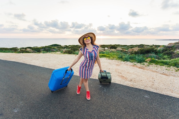 Счастливый путешественник женщина сидит на чемодане на дороге и улыбается. Концепция путешествия, путешествия, поездки.
