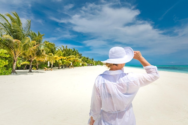 하얀 드레스를 입은 행복한 여행자 여성은 열대 해변 휴가를 즐기고 바다 해안을 바라보고 있습니다.