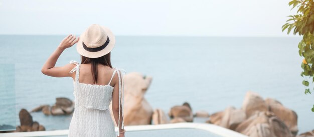 Happy traveler woman in white dress and hat enjoy beautiful sea view young woman standing at swimming pool Freedom relaxing vacation holiday and summer travel concept