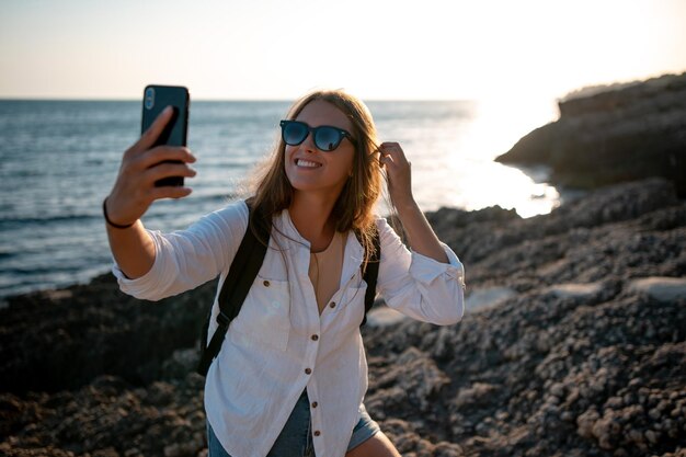 Happy traveler woman in white shirt and sungalsses taking selfie with mobile phone on the rocky shore at beautiful sunset