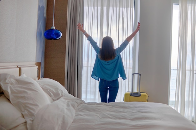 Happy traveler woman with raised hands and suitcase standing near window and looking to a view from hotel room after check-in