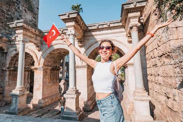 トルコの旗を手に持ち、アンタルヤの有名な門またはハドリアヌスのアーチを持つ幸せな旅行者の女の子