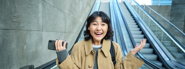 Happy triumphing asian girl looks amazed and joyful holds smartphone goes down escalator with