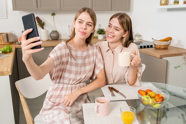 Happy twin teenage girls making selfie or video calling in the kitchen