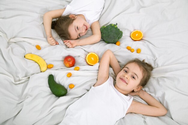 幸せな2人のかわいい子供たちは、ベッドの上で果物や野菜で遊んでいます。子供のための健康食品。