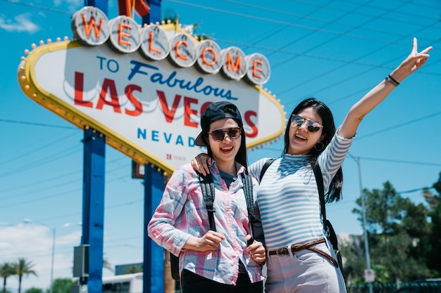 happy two cute young asian women travelers giving V signs as they stand arm in arm grinning at camera in urban on sunny summer day. sisters tourists taking picture with las vegas sign together