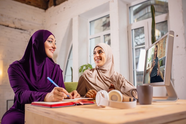 Happy two muslim women at home during lesson, studying near computer, online education