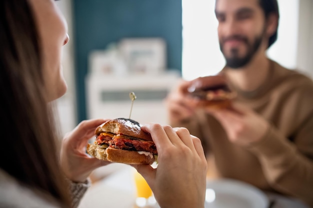 Happy unrecognizable young couple in love eating hamburgers indoors at home.