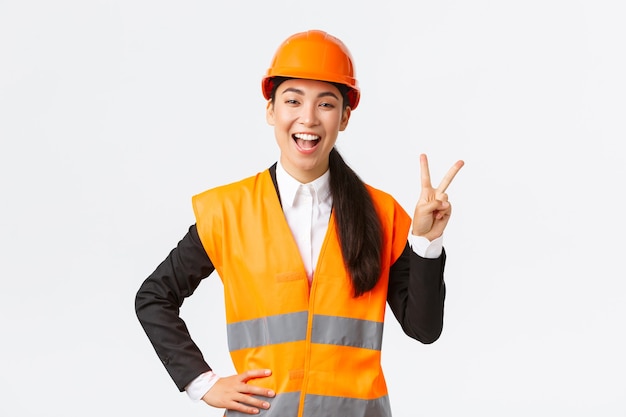 Happy upbeat asian female asian architect in safety helmet and reflective jacket, showing peace sign and smiling, assured in victory of her company, winning tender on construction works