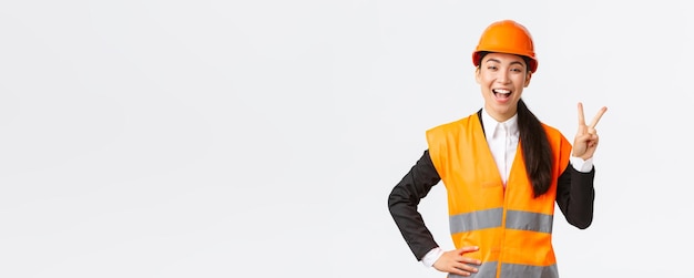 Happy upbeat asian female asian architect in safety helmet and reflective jacket showing peace sign and smiling assured in victory of her company winning tender on construction works