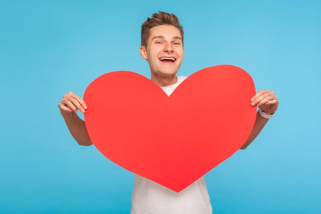 Happy Valentine day. Portrait of delighted cheerful man holding big red heart and smiling broadly to camera, declaration of love, romantic present. indoor studio shot isolated on blue background