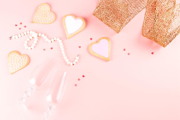 Поздравительная открытка на день Святого Валентина с сердечным печеньем, бокалами и вином на пастельно-розовом фоне.