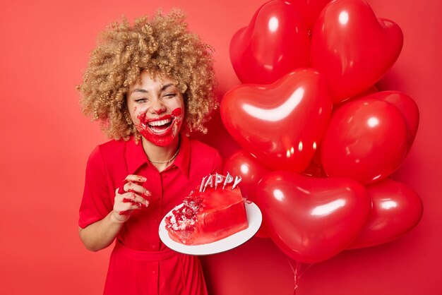 Happy Valentines Day concept Vrolijke krullend haired vrouw besmeurd met lekkere cake crème geniet van viering heeft romantische date met vriendje draagt jurk poses in de buurt van stelletje hartvormige ballonnen