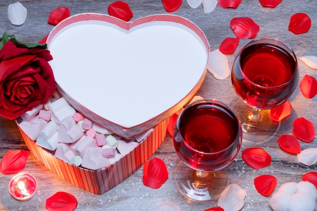 Happy Valentines Day gift box in the shape of a heart and two glasses of red wine rose petals lie