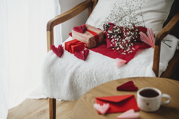 Happy valentines day Modern gifts red envelope and white flowers on cozy armchair on background of defocused stylish cup of tea with heart on wooden table Valentine morning surprise