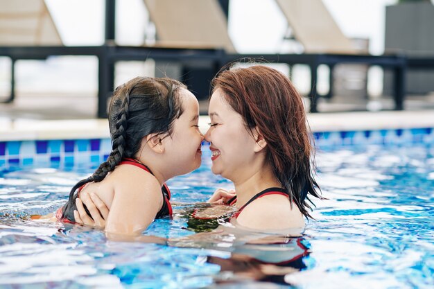 Happy Vietnamese mother and daughter touching noses when standing in swimming pool