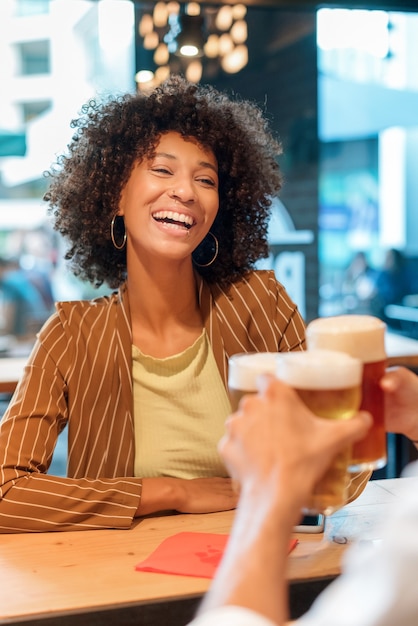 Foto felice vivace ridere giovane donna nera brindare con una birra fredda mentre festeggia con un amico in un pub o ristorante in una strada cittadina