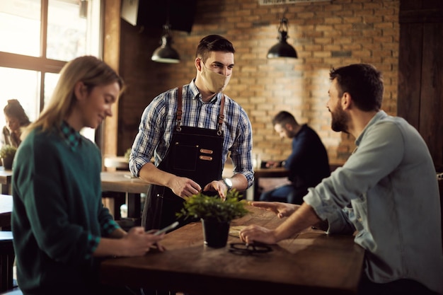 Happy waiter wearing face mask while talking to customers n a pub