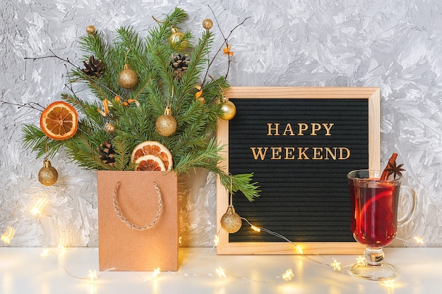 Happy Weekend text on black letter board, festive decorated christmas tree in craft package, glass of hot mulled wine 