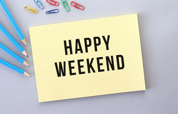 HAPPY WEEKEND text in notebook on gray background next to pencils and paper clips.