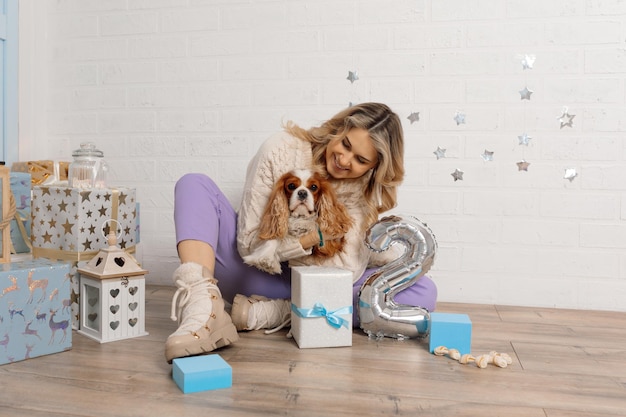 Счастливая ухоженная женщина обнимает собаку спаниеля, крепко сидящего на полу рядом с подарочными коробками и серебряным номером два