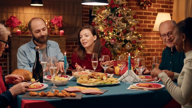 Happy wife talking to guests about food while sitting together at Christmas dinner table. Festive cheerful people enjoying traditional winter holiday meal while celebrating feast.