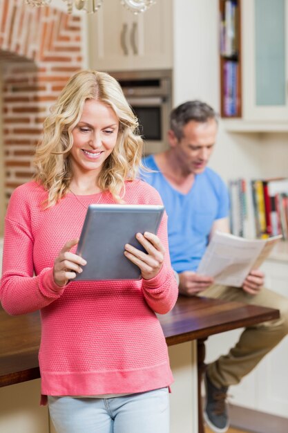 Happy wife using tablet while husband reading newspaper in the kitchen