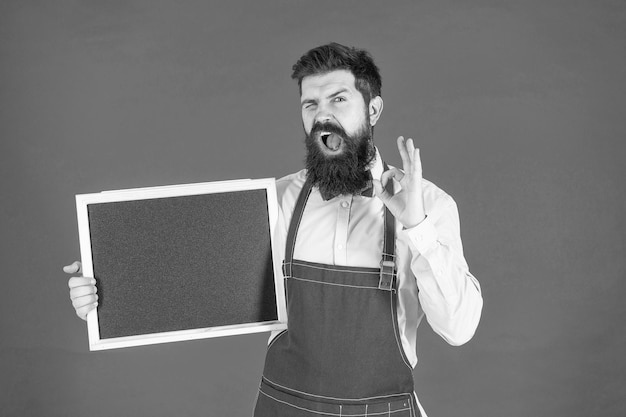 Happy winking man in barber apron hold school blackboard showing OK sing ring gesture red background copy space enrollment