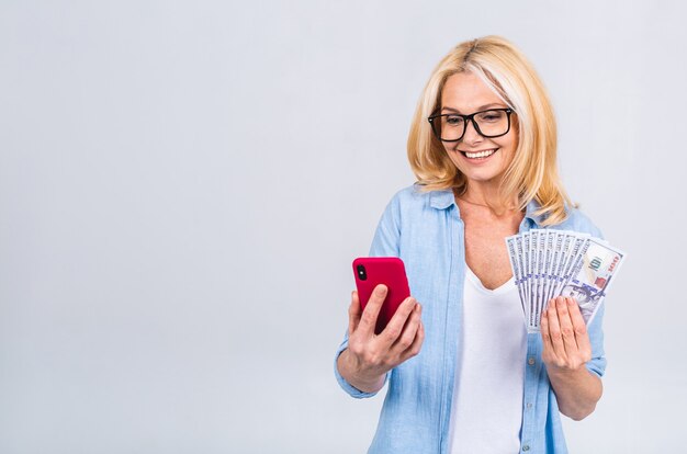 Happy winner! image of mature happy senior woman standing isolated over white background wall looking at camera holding money using mobile phone