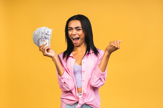 Happy winner portrait of african american successful woman 20s with afro hairstyle holding lots of money dollar banknotes isolated over yellow background