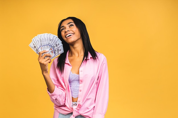 Happy winner Portrait of african american successful woman 20s with afro hairstyle holding lots of money dollar banknotes isolated over yellow background