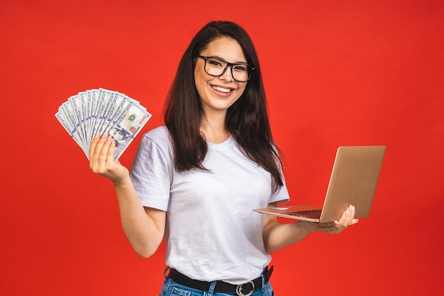 Happy winner Pretty young business woman in casual holding laptop in the office isolated over red background Working with computer holding money bills