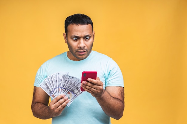 Happy winner! Young rich african american indian black man in casual holding money dollar bills and mobile phone with surprise isolated over yellow background.