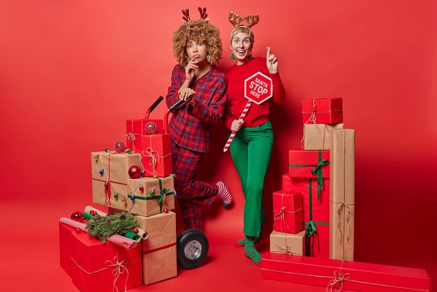 Happy winter holidays concept two women stand next to each
other keep fingers crossed make wish stand around pile of gift
boxes prepare for christmas celebration stand in full length
against red wall