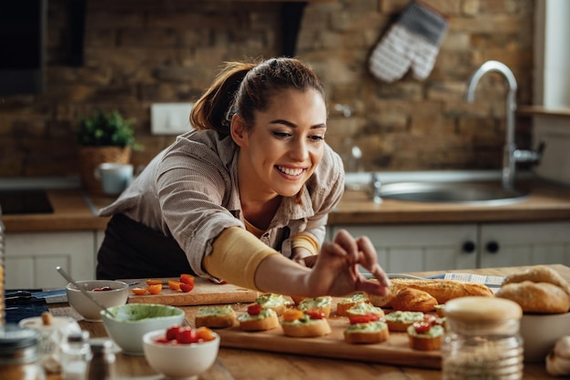 Foto donna felice che aggiunge pomodoro ciliegino mentre prepara la bruschetta in cucina.